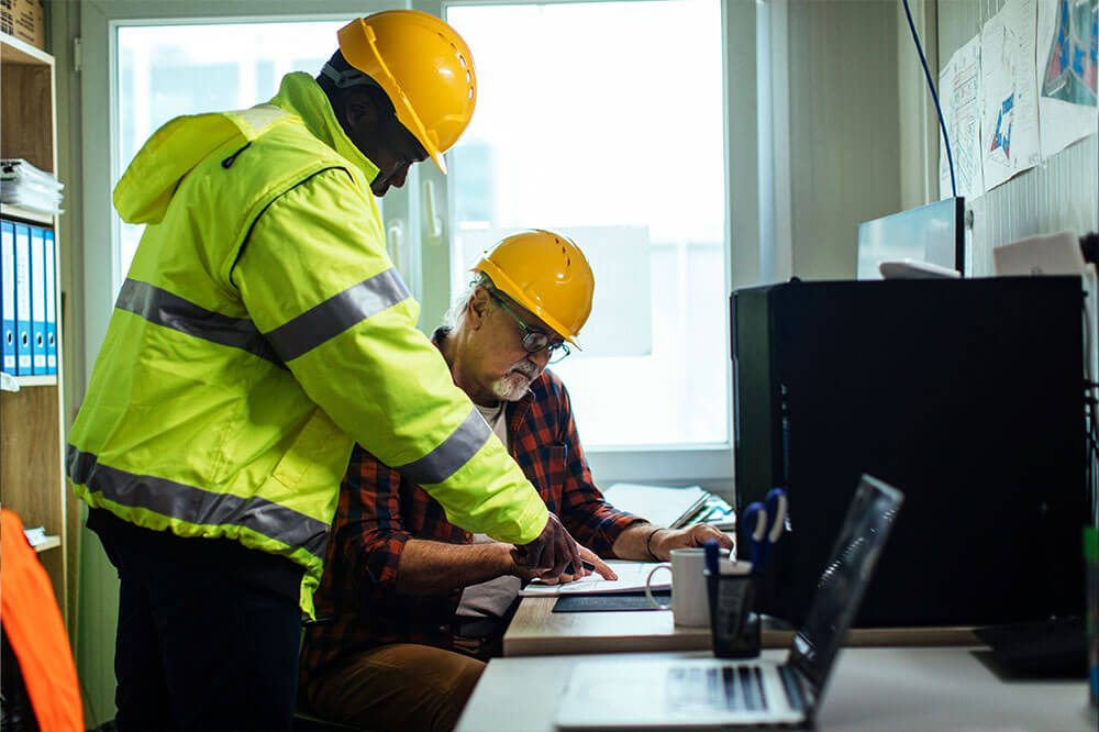 two men in hardhats inside of an office looking at data